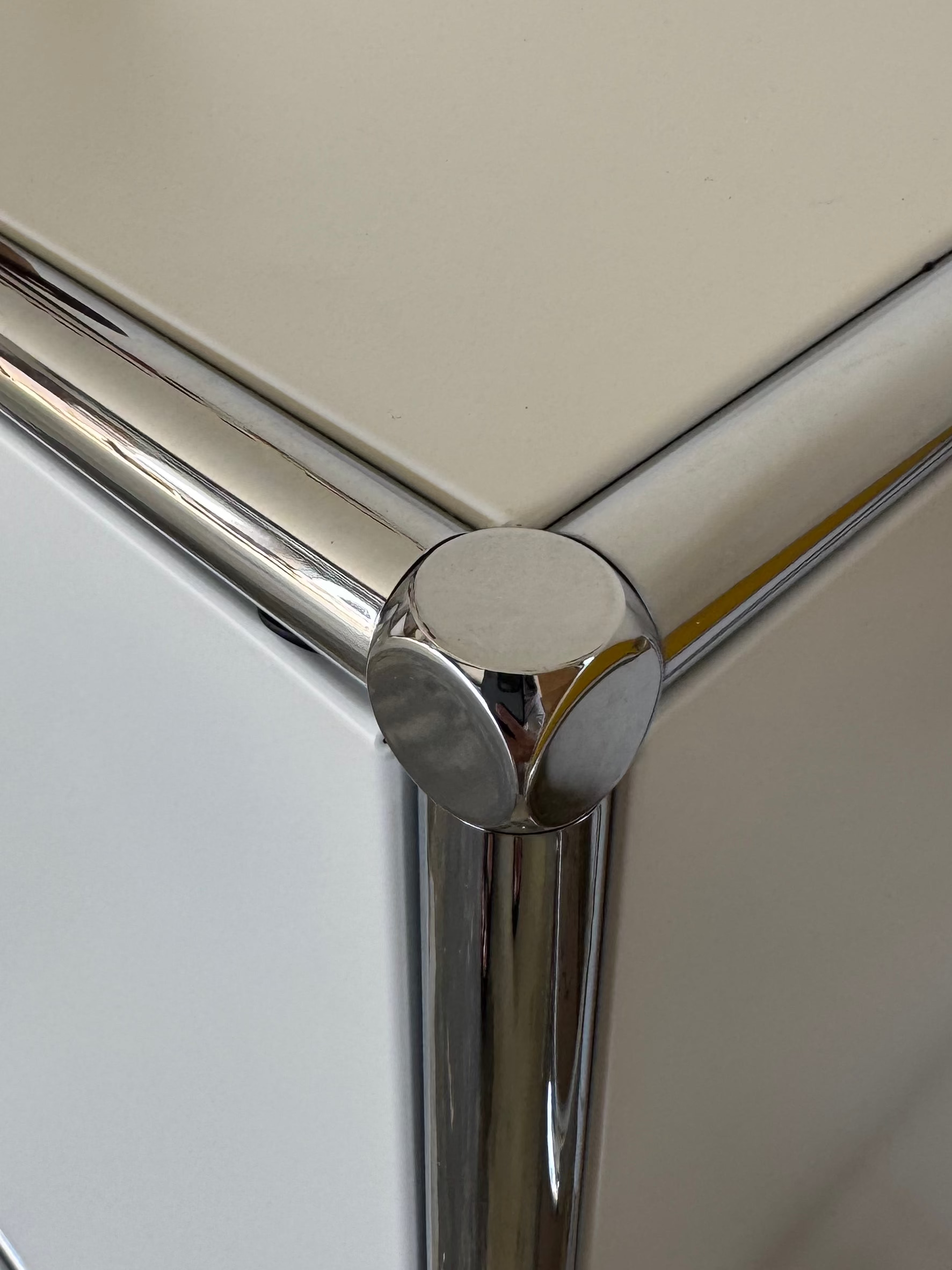 A close-up of a white cabinet or dresser corner with shiny chrome metal trim and protective round corner guards, with a mushroom-shaped decorative object on top.