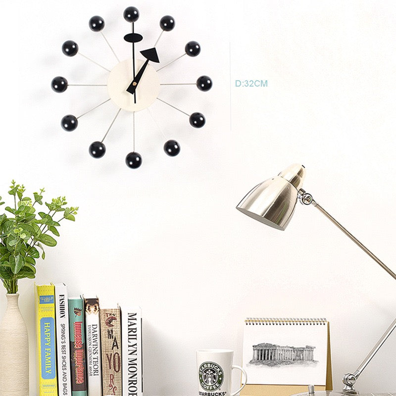 Modern wall clock with black and white design on a white wall, with books and a lamp in the foreground.