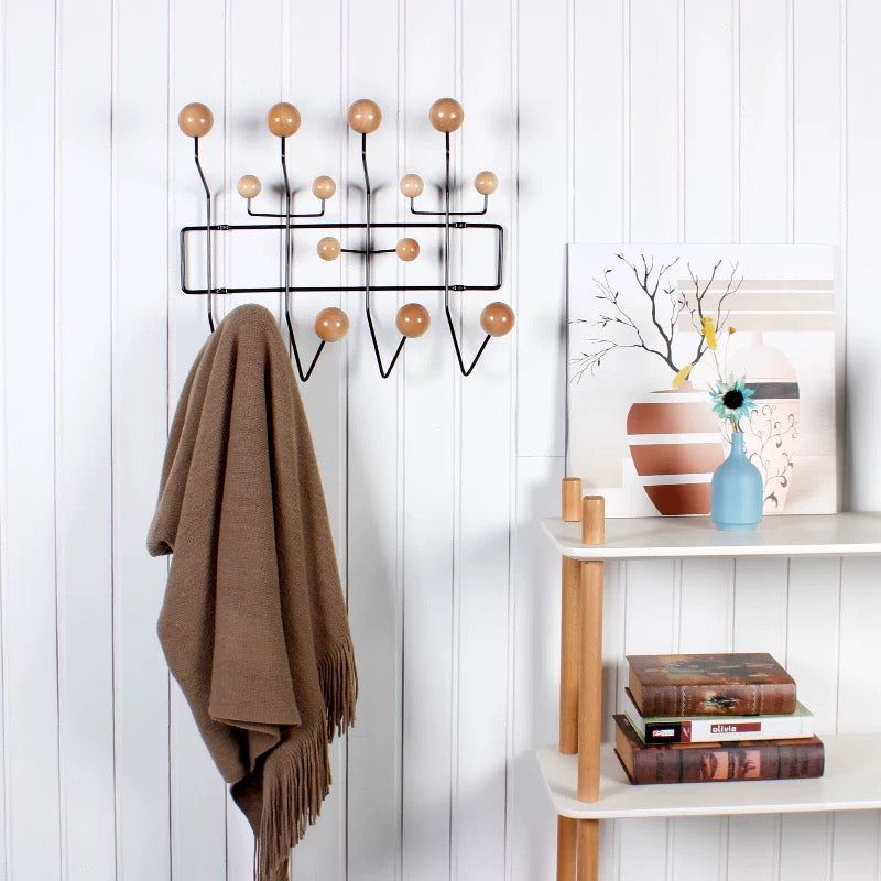 Wall-mounted coat rack with wooden hooks on a white paneled wall, next to a shelf with books and a vase.